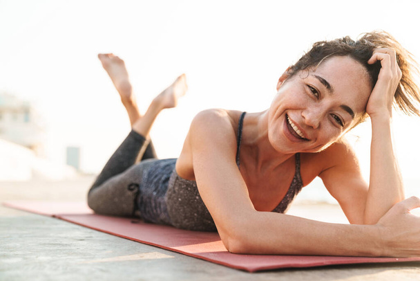 Photo of joyful sportswoman in tracksuit smiling and lying on fitness mat while practicing yoga in morning outdoors - Fotó, kép