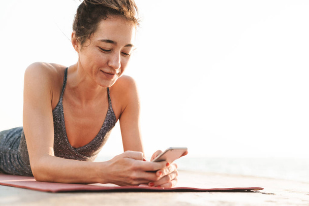 Photo of caucasian sportswoman in tracksuit holding smartphone and lying on fitness mat while practicing yoga by seaside in morning - Fotoğraf, Görsel