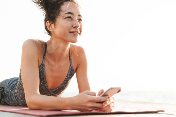 Photo of happy sportswoman in tracksuit holding smartphone and lying on fitness mat while practicing yoga by seaside in morning - Foto, Imagem