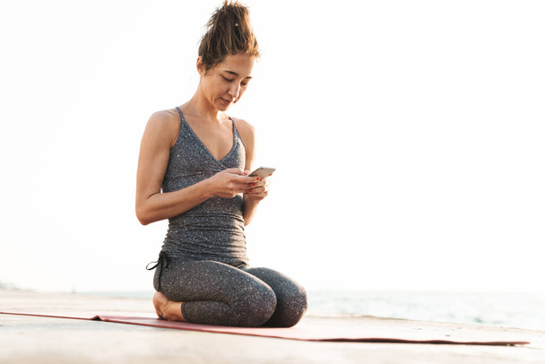 Photo of healthy sportswoman in tracksuit holding smartphone and sitting on fitness mat while practicing yoga by seaside in morning - Foto, immagini