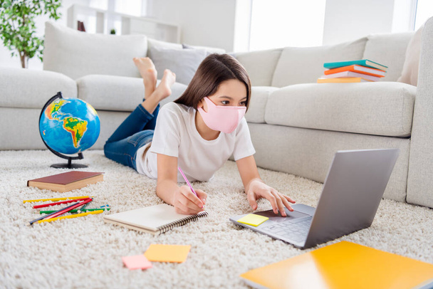 Photo of small pupil school girl search information notebook homework write notes geography interesting facts lying floor domestic social distance quarantine study safety face mask indoors - 写真・画像