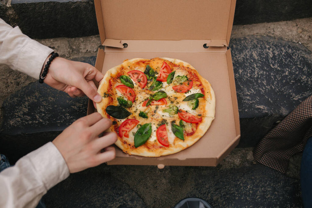 Man grab slices of pizza from box at the outdoor. Male hands taking pizza. Vegan pizza with fresh tomatoes basil and broccoli. Lactose and gluten free - Foto, imagen