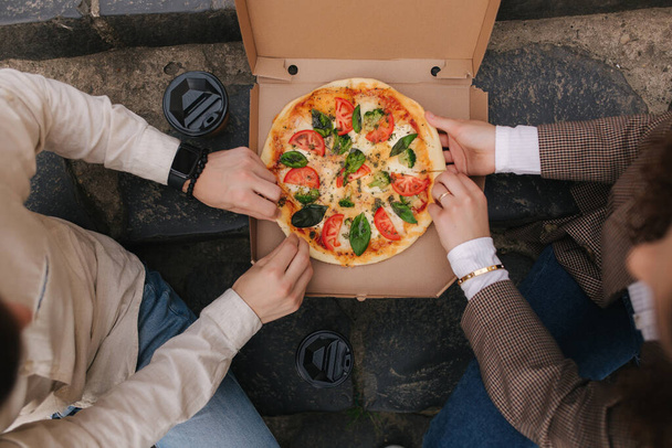 Widok z góry obraz pary chwycić plasterki pizzy z pudełka na zewnątrz. Mężczyzna i kobieta biorą pizzę. Wegańska pizza ze świeżymi pomidorami bazylia i brokuły. Laktoza i gluten bez - Zdjęcie, obraz