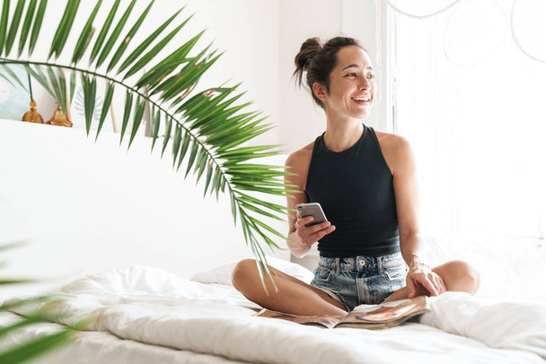 Portrait of satisfied woman in summer clothes holding mobile phone while reading magazine on bed in bright room with green plant - Valokuva, kuva
