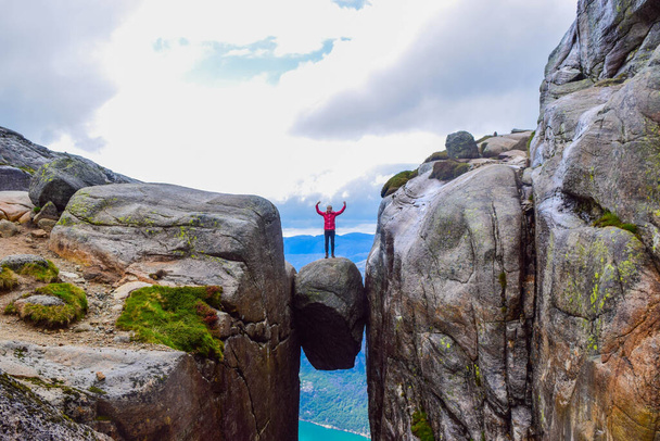 The tourist hiker girl standing on top of Kjeragbolten - the most dangerous stone in the world. Kjeragbolten is a rock stuck at an altitude of 984 meters above Lysefjorden on mountain Kjerag, Norway. - Fotó, kép