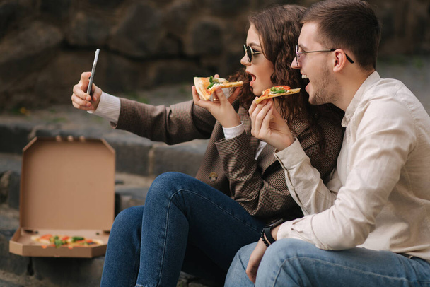若いブロガーは手にピザのスライスで自撮りを行います.カメラの電話にサングラスの笑顔で美しいカップル。ビーガンファーストフード - 写真・画像