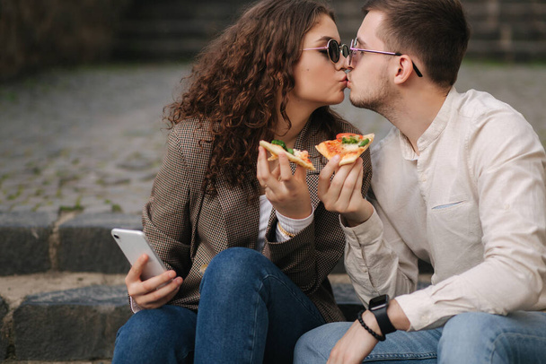 Young bloggers make selfie with slice of pizza in hands. Beautiful couple in sunglasses smile and kiss. Vegan fast food - Photo, Image