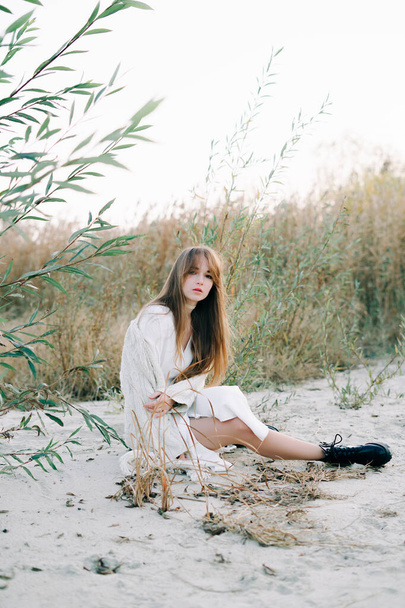 portrait of a young girl in a white dress, a warm knitted cardigan and black shoes among dry reeds and green bushes on the Bank of a river with white sand - Foto, imagen