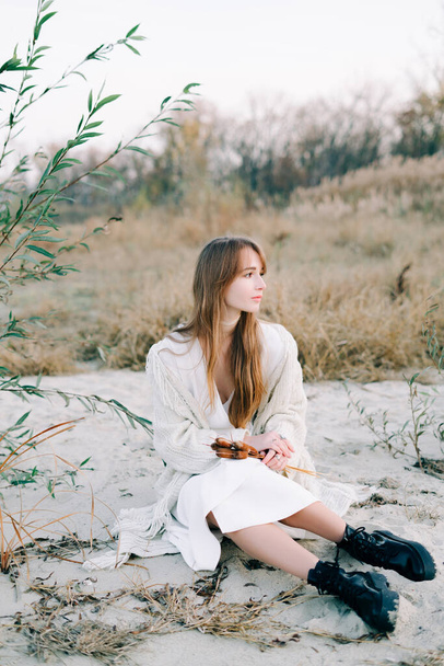 portrait of a young girl in a white dress, a warm knitted cardigan and black shoes among dry reeds and green bushes on the Bank of a river with white sand - Photo, Image