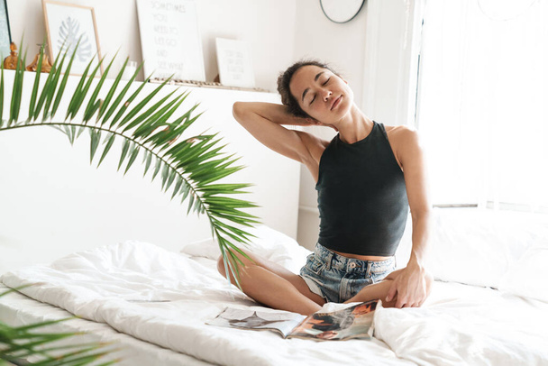 Portrait of relaxed young woman in summer clothes reading magazine on bed in bright room with green plant - Foto, imagen