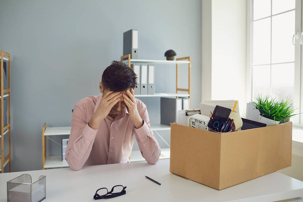 Unemployment. Dismissal. Dismissed businessman is upset with a cardboard box holding his head in his hands while sitting in a company office. - Foto, imagen