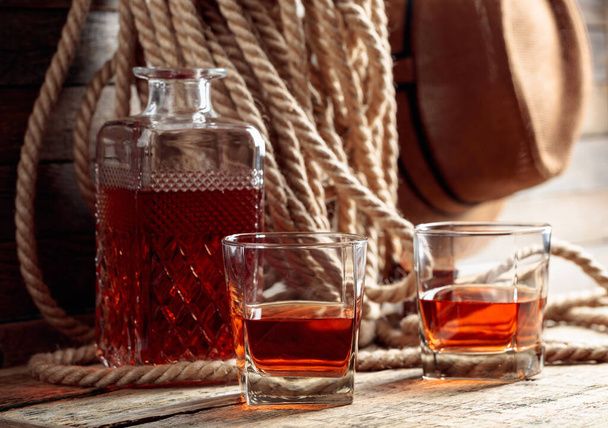 Glas und Karaffe mit starkem Alkohol auf einem alten Holztisch. Im Hintergrund ein Hanfseil und ein Männerhut.  - Foto, Bild