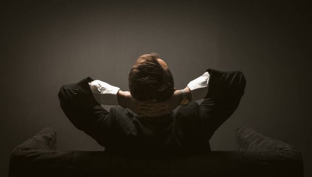 Businessman pondering new startup project sitting on a sofa. Rear view of Man in black suit sitting at couch in dark room while thinking with hands behind his head - Foto, Bild