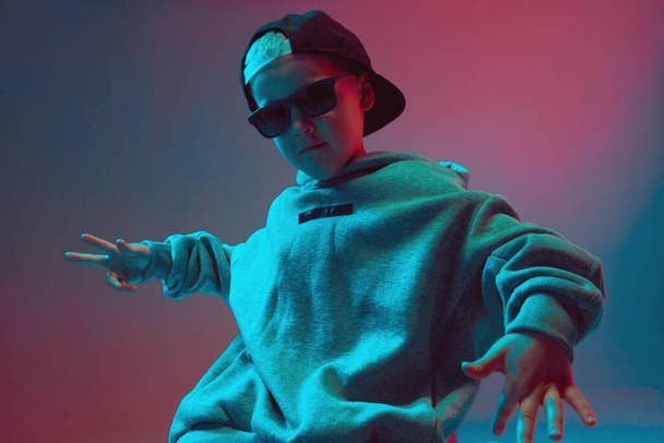 Portrait of a cool boy child in a rap image, stylishly posing in a hoodie, sunglasses and a cap on a neon background. - Foto, Imagen