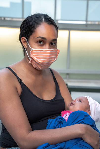 Een vrouwelijke gemengde ras Afro-Amerikaanse moeder in een zwarte tank top draagt een geïmproviseerde gestreepte oranje stof masker en houdt haar baby baby meisje verpakt in haar blauwe jas tijdens de COVID-19 pandemie. - Foto, afbeelding