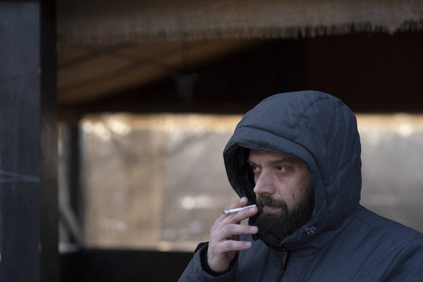 Πορτρέτο του άντρα με την κουκούλα και το τσιγάρο. Ένας άντρας με γενειάδα σε γκρι σακάκι στο δρόμο. Το πορτρέτο ενός άντρα. Ο τύπος με το μούσι. Χειμώνας, χειμωνιάτικο μπουφάν, γένια. Πορτραίτο ενός άντρα με κουκούλα - Φωτογραφία, εικόνα