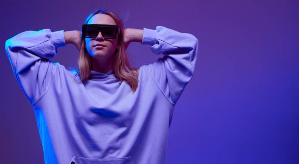 Portrait of a stylish young girl, cool posing in a hoodie, sunglasses and with developing hair, on a neon background. - Фото, зображення