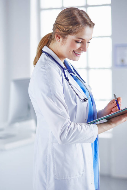 Giovane donna medico è in piedi con bordo con appunti sorridente in ufficio ospedaliero
 - Foto, immagini