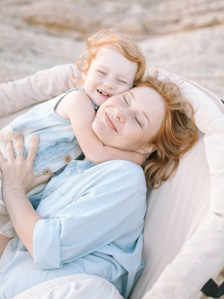 αξιολάτρευτο υπέροχο παιδί και η μητέρα της με κόκκινα μαλλιά ντυμένοι με γαλάζιο πουκάμισο, αγκαλιάζονται και γελάνε. - Φωτογραφία, εικόνα