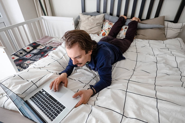 Ο άνθρωπος που προσπαθεί να εργαστεί μέσω φορητού υπολογιστή στο σπίτι κατά τη διάρκεια της καραντίνας με το αγοράκι να πηδάει στο κρεβάτι. Οικογενειακή ζωή κατά τη διάρκεια της απομόνωσης. Μείνε σπίτι. Εργασία από το σπίτι, γραφείο στο σπίτι - Φωτογραφία, εικόνα