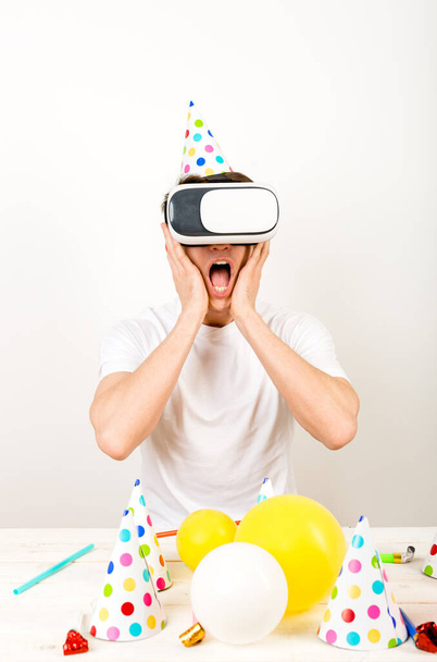 A guy wearing VR helmet sitting on a white wall in front of a deck full of party stuff and shows strong expressions  - Photo, Image