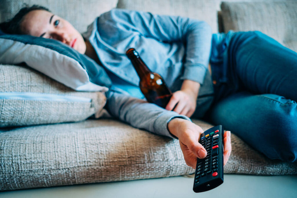 The girl lies on the couch with a bottle of alcohol and a remote control from the TV. Close up. - Photo, Image