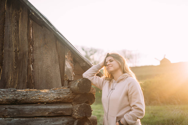 Zamazana dziewczyna w delikatnym różowym swetrze stoi obok drewnianego starego domu i uśmiecha się - Zdjęcie, obraz