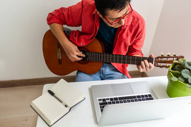 W pokoju w domu mężczyzna gra na gitarze przed laptopem podczas zajęć online. - Zdjęcie, obraz