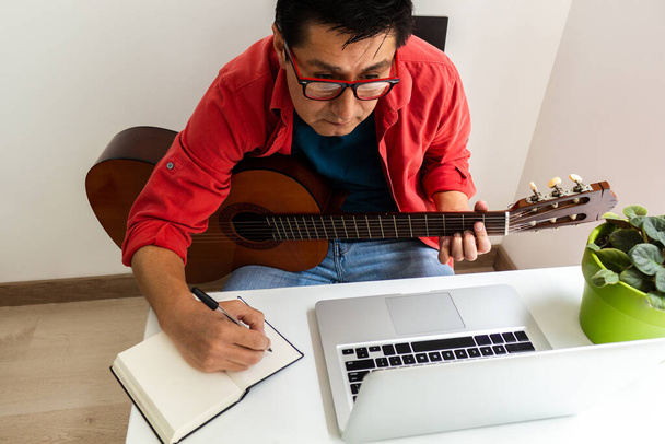Un uomo che riceve lezioni di chitarra sul suo portatile a casa. Prende appunti in un quaderno. Attività durante la quarantena
 - Foto, immagini