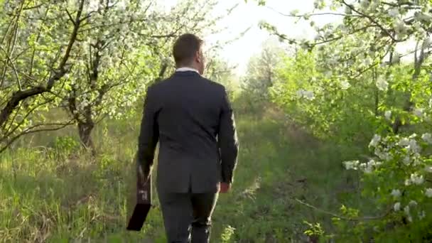 Een zakenman loopt door de appelboomgaard en kijkt rond. Een man in een pak kijkt naar zijn bezittingen. Een man in de natuur. - Video