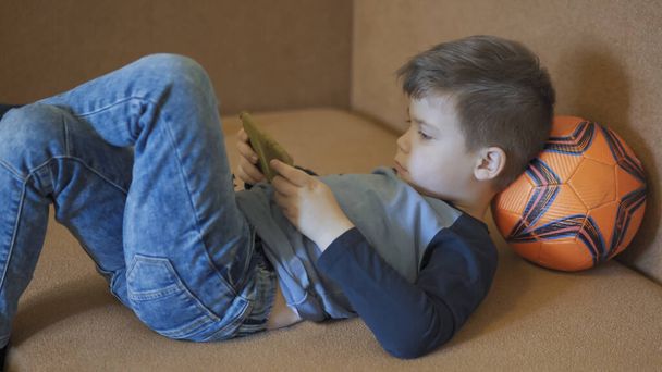 Preschooler looks at mobile phone screen lying on sofa with head resting on soccer ball. Boy studying online because of quarantine. Home education concept. Profile view - Foto, Imagen