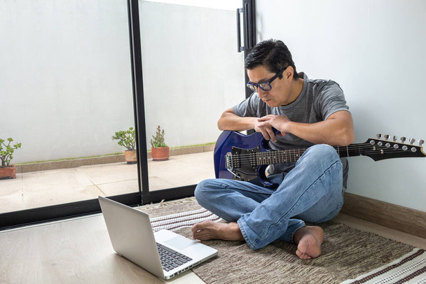 Durante la quarantena un uomo guarda una classe di chitarra elettrica online, è seduto su un tappeto in una stanza a casa - Foto, immagini