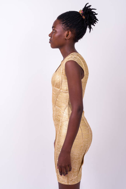 Στούντιο πλάνο της νεαρής όμορφης Αφρικής γυναίκα φορώντας χρυσό φόρεμα έτοιμο να κόμμα σε λευκό φόντο - Φωτογραφία, εικόνα