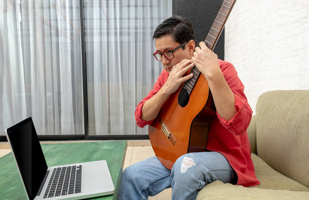 En el salón de su casa, un hombre mira de cerca a su ordenador recibiendo una clase de guitarra virtual. - Foto, imagen