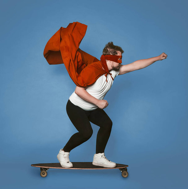 Super speed concept. Superhero rushes forward on a skateboard. Man in red superhero costume with a flying cloak isolated on faded denim blue background - 写真・画像