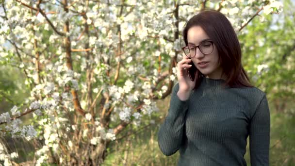 Jonge vrouw in de natuur met een telefoon. Een meisje praat aan de telefoon terwijl ze tegen een bloeiende boom staat. - Video
