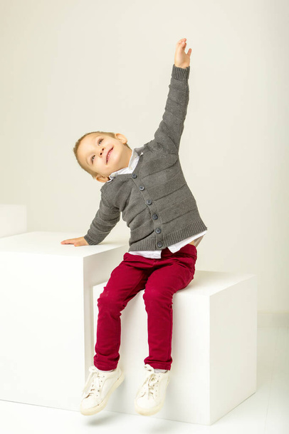 Roztomilý šťastný usměvavý chlapec sedí na bílé kostce ve studiu - Fotografie, Obrázek