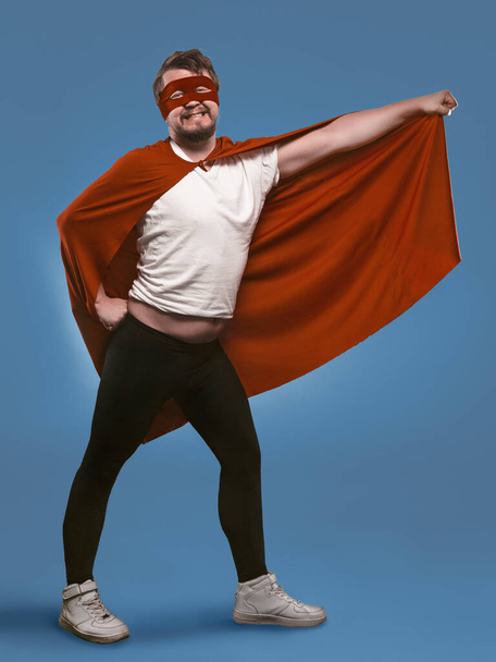 Smiling super hero man ready to fly save world. Funny man in red superhero costume and mask holding his cloak with hand posing in full growth on faded denim blue background - Foto, Bild