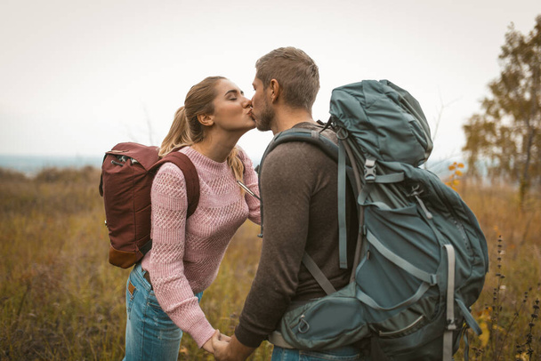 Loving backpackers kiss in the field outdoors. Happy couple of travelers, young man and woman standing against the backdrop of autumn nature. Love for adventure concept - Valokuva, kuva