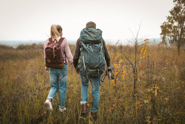 Hiking in autumn nature, couple of backpackers makes their way across the field, rear view of man and woman with backpacks and hiking poles walking in nature outdoors. Hiking concept - Фото, изображение