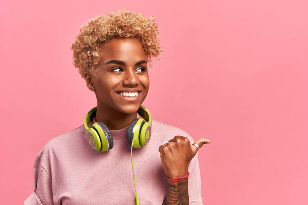 African American woman in high spirit points away with thumb, feels entertained and satisfied, listens radio broadcasting with modern headphones connected to telephone, checks sound, shows free space - Foto, Imagen