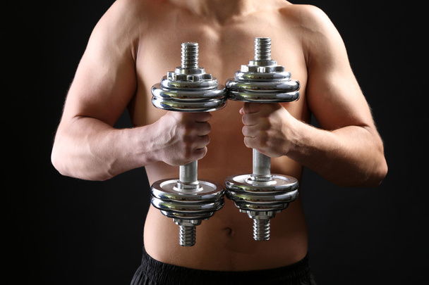 Handsome young muscular sportsman execute exercise with dumbbells on dark background - 写真・画像