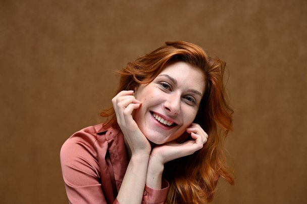 Photo portrait on a beige background of a happy pretty young woman in a pink shirt with long beautiful red hair. Studio shot. - Zdjęcie, obraz