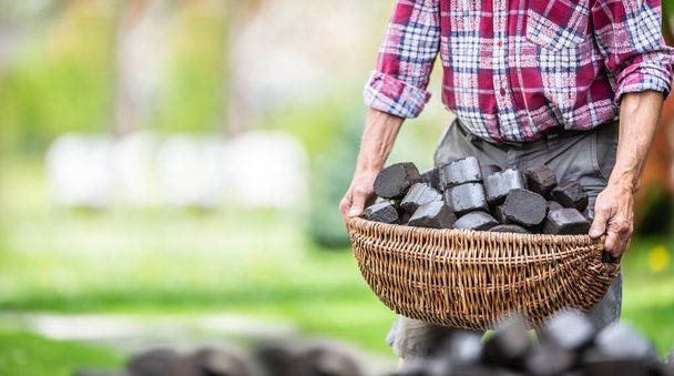 Человек несет корзину, полную угольных брикетов на заднем дворе своего дома
. - Фото, изображение