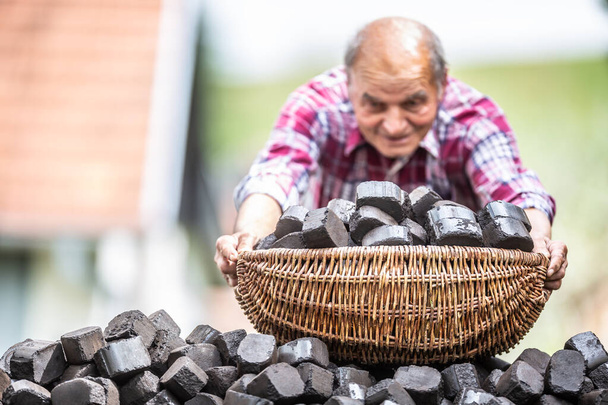 Old man picking up a basket full of coal briquettes from a pile in the backyard. - Photo, image