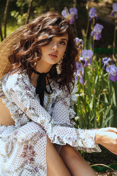 fashion outdoor photo of beautiful woman with dark curly hair in elegant dress and straw hat posing in summer garden - 写真・画像