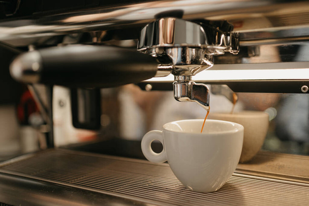 Kahve dükkanında profesyonel bir espresso makinesinde Espresso demlemek. Kafedeki kahve makinesinden beyaz bir fincana dökülen kahvenin yakın çekimi.. - Fotoğraf, Görsel