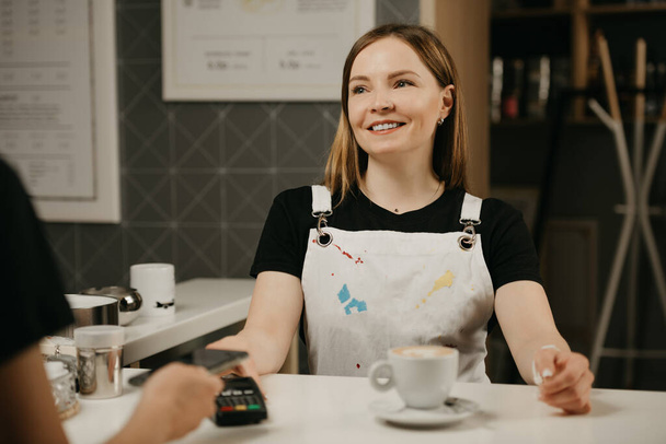 A close portrait of a female barista who smiling holds out to a client a terminal for paying. A woman paying for a cup of latte with a smartphone by contactless PAY PASS technology in a cafe.  - Φωτογραφία, εικόνα