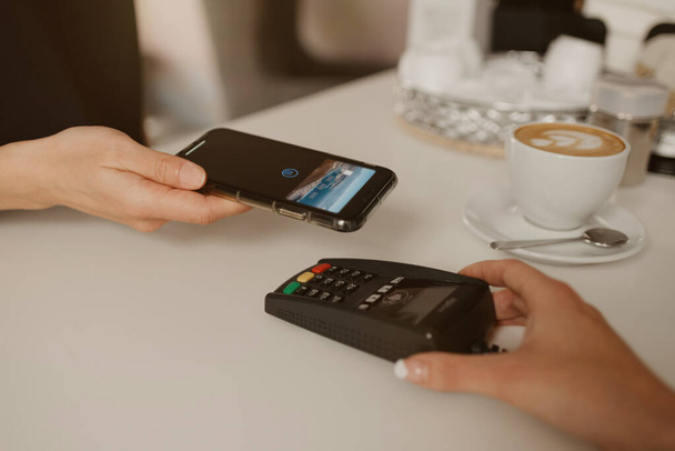 Nainen maksaa lattensa älypuhelimella kontaktittomalla NFC-tekniikalla kahvilassa. Naispuolinen barista tarjoaa terminaalin asiakkaalle maksamiseen kahvilassa
. - Valokuva, kuva
