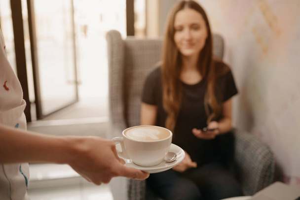 Detailní záběr na latté, které baristka podrží dívce v kavárně. Žena s dlouhými vlasy pracující na dálku na notebooku udržuje společenskou vzdálenost uchopí šálek kávy v kavárně.   - Fotografie, Obrázek